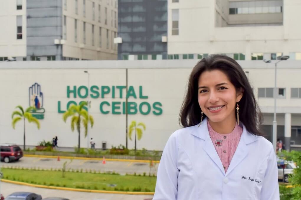 Medical professional standing outside a hospital in Ecuador, wearing a white lab coat and smiling toward the camera.